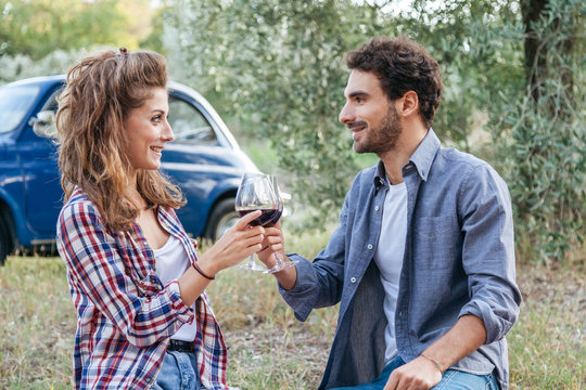 Couple of young lovers drinking red wine from glass goblet at picnic in countryside, Tuscany, Italy. Sitting on lawn among the olive trees on a late summer day, behind them  old blue car