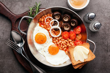 Breakfast with fried eggs in cast-iron pan, broken egg, tomatoes, on cutting board over grey stone background. Top view with copy space