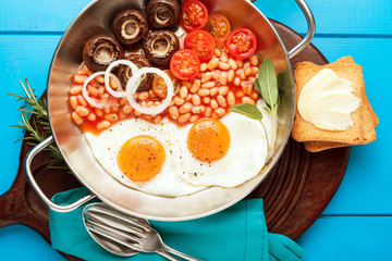 Breakfast with fried eggs in cast-iron pan, broken egg, tomatoes, on cutting board over turquoise wooden background. Top view with copy space