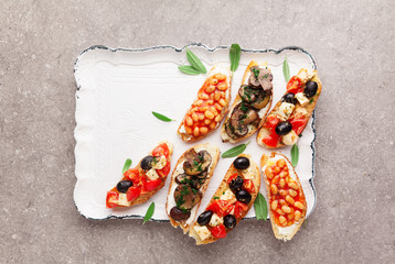 Variety of small bruschetta with caprese salad , white beans on the grill with tomato sauce and fried mushrooms with soft cheese, served on grey  background.