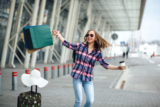 Attractive hipster woman in sunglasses dancing and whirling in airport with disposable cup of coffee, paperbags, suitcase and hat and at gate waiting in terminal.