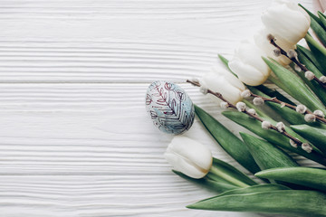 happy easter greeting card. stylish easter egg and willow buds and white tulips on rustic wooden background flat lay. concept with space for text, top view. soft light