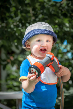 Little cute child in blue hat holding a garden hose outdoors sunny summer day, selective focus.