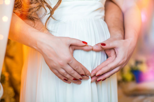 Pregnant girl in white dress, her hands, and husband's hand on her stomach in form of heart