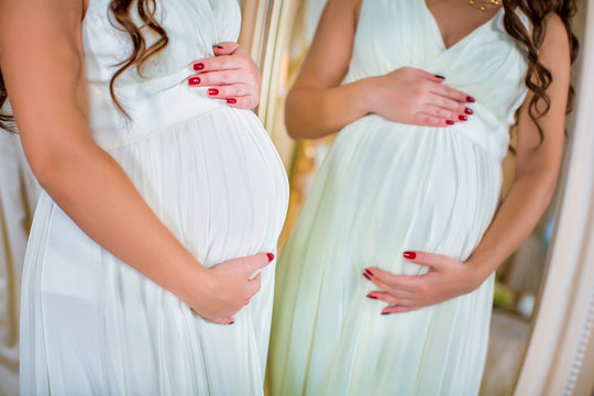 two pregnant women in white dresses, their hands on her stomach