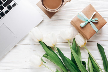 Obraz na płótnie Canvas laptop with morning coffee and tulips and stylish gift box on white wooden rustic background. flat lay freelance, work gadgets with space for text. spring holiday. happy women day
