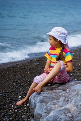  A child girl sitting on the beach on a rocky beach and looks into the distance.