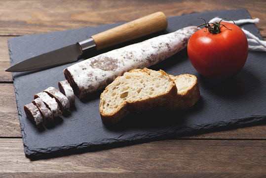 Fuet and a knife next to a tomato on a slate plate. Spanish sausage. Horizontal shoot.