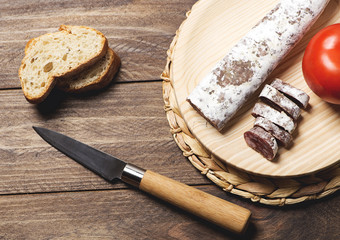 Fuet bread and a knife next to a tomato on wooden table. Spanish sausage. Horizontal shoot.