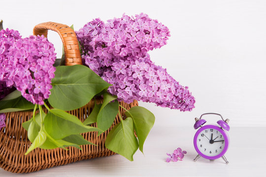 Bouquet of lilac in wicker basket with purple alarm clock on white background