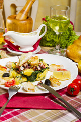 Grilled calamari on a white plate with boiled potatoes and chard, lemon, cherry tomatoes and olives, served next to sauce and glass of white wine.