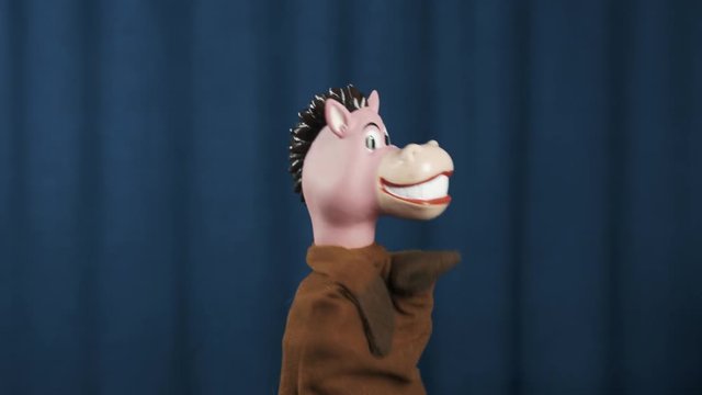 Smiling horse hand puppet toy nodding head and shaking arms wide spread on scene with blue crease curtains background
