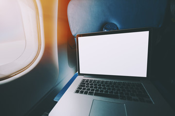 Wide angle shoot of modern laptop with blank screen on the blue table of airplane seat, mock up of portable computer next to porthole of flying plane with sunny day outside