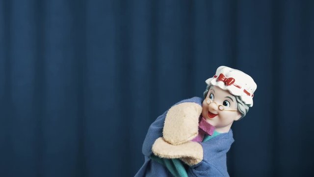 Elder woman hand puppet makes tossing movements and brake dances on blue crease curtains background on scene