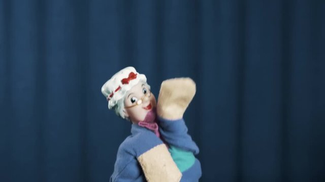 Old woman hand puppet appears on scene with smooth dance movements on blue crease curtains background on scene