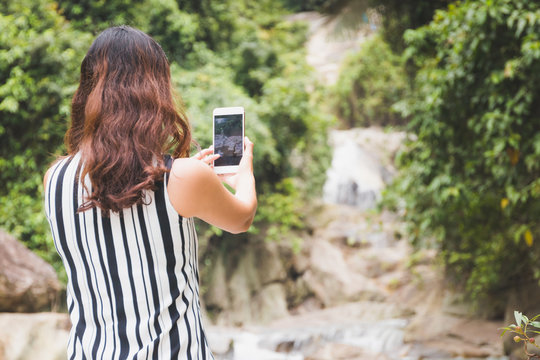 Asian woman tourist taking photo with camera phone at waterfall in Thailand