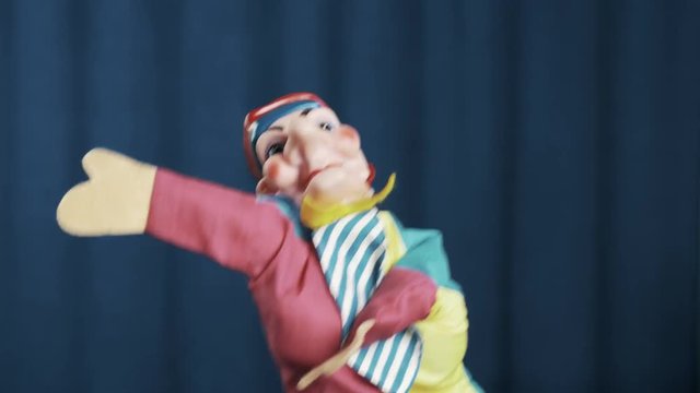 Funny jester hand puppet toy appears on scene with blue crease curtains background, greeting audience