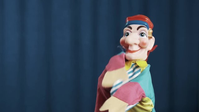 Funny jester hand puppet toy dancing and spread arms on scene with blue crease curtains background