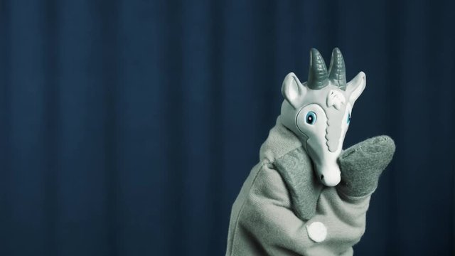 Gray blue eyed goat hand puppet toy appears on scene with blue crease curtains background, greeting audience and telling story
