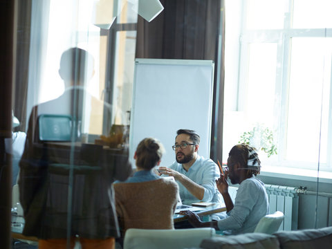 Group Of Creative Young People, Afro-American Man Among Them, Sitting Round Meeting Table In Modern Conference Room Listening Intently To Asian Speaker, Shot From Behind Glass Wall