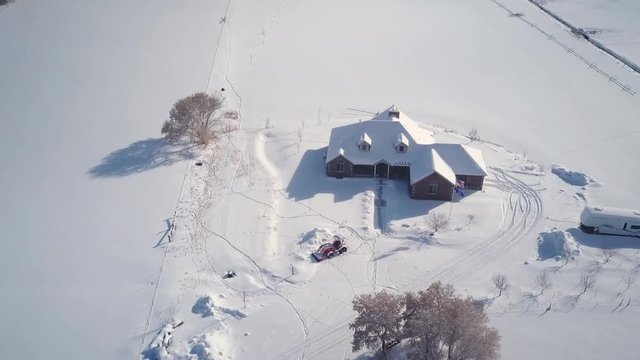 Aerial farm house winter snow tractor working fast motion. Aerial point of view. Winter storm and blizzard drops fresh white snow. Seasonal rural farm community. Frigid season weather hazard.