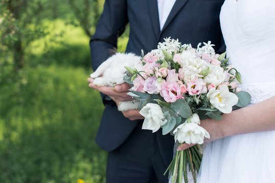 Wedding day. Bride holding in her hands a delicate wedding bouquet with white and pink tulips and pink small roses. Groom holding a white cute rabbit. 