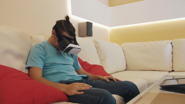 Young caucasian naughty man wears virtual reality headset in blue shirt with hair knot sitting on white leather sofa in modern apartment living room watching lewd video game.