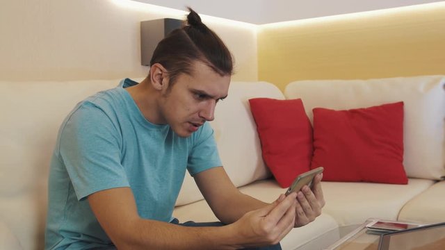 Young Caucasian Cheerful Man In Blue Shirt With Hair Knot Sitting On White Leather Sofa In Modern Apartment Living Room Talking Video Call On Smartphone