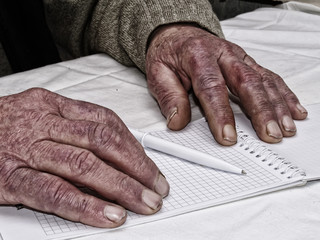 Closeup of the wrinkled hands of an old caucasian man holding pen and paper, wearing a green sweater