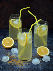 Three glasses of cold homemade lemonade with lemon slices, ice cubes, brown sugar and straws on dark background.