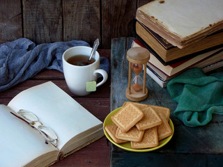 The composition of a stack of old books, open book, tea cups, glasses and plates of sugar cookies on a wooden background. Vintage photo.