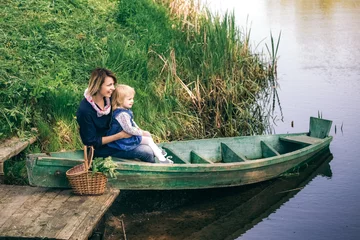 Foto op Canvas Portrait of happy family of two people on vacation. Young mother and little daughter on picnic sitting in old wooden boat on spring river over water background. Age of child 2 years and 4 month. © Andrii Oleksiienko
