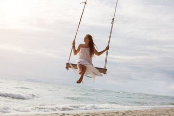 Beautiful young female sitting on the swing on sea shore enjoy summer - obrazy, fototapety, plakaty