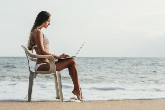 Young woman in white swimsuit using laptop with internet sitting on a chair on tropical island beach. Summer travel, freelance concept, shopping online