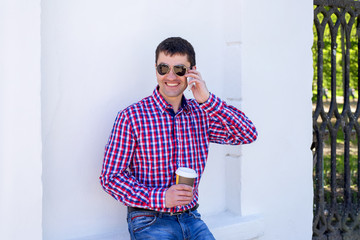 Man against a wall, with coffee talking on the phone, in the summer with glasses and shirt, happy smiling businessman.