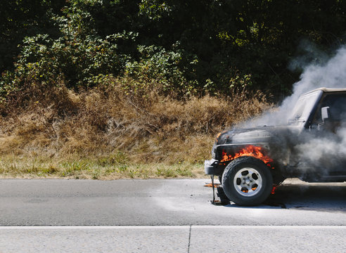 A burning car parked on a country road in front of bushes and trees.