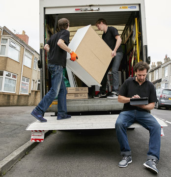 Removals business. A removals company, two men lifting furniture and one seated using a digital tablet. 