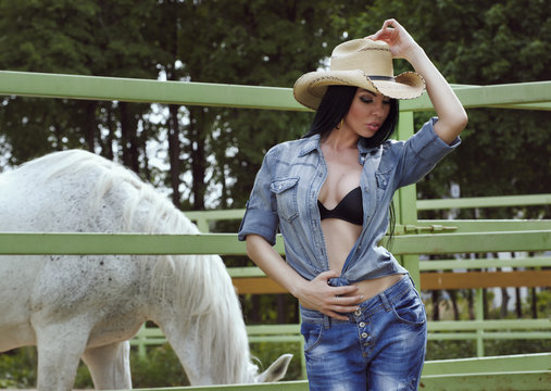 Beautiful cowgirl in hat and jeans near a corral with a horse