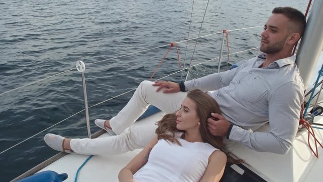 Beautiful woman lying on lap of her boyfriend on deck of yacht sailing on lake. They looking far away and man stroking hair of girlfriend