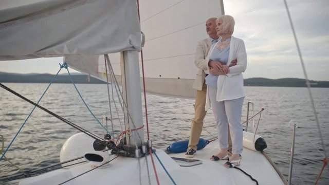 Senior man and woman standing at deck of drifting yacht, talking and embracing