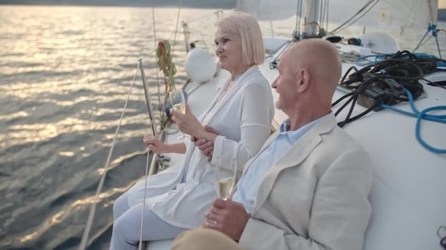 Joyous senior couple sitting aboard yacht with champagne glasses, talking and laughing