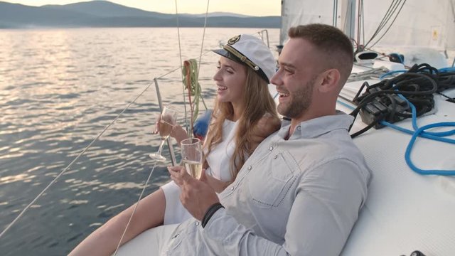 Joyous young woman in sailor cap sitting aboard with her boyfriend. They holding champagne glasses, laughing and enjoying panoramic view of lake