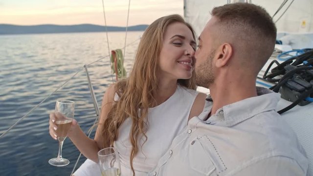 Closeup of young boyfriend and girlfriend sitting on edge of yacht with glasses of wine, rubbing noses and kissing