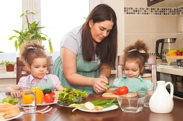 Family preparing meal together. Happy mother and her cute twin daughters having fun cooking vegetable salad.
