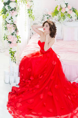 Beautiful woman in red long dress with a gold crown earrings and posing while sitting on a bed