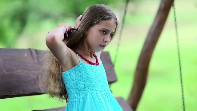 Attractive girl in blue sundress sits on the bench and combs his long hair. Girl with hairbrush sits on bench in garden. Beautiful girl combs his long hair