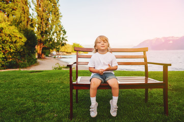 Cute toddler boy playing outdoors, resting on the bench by Lake Geneva on a nice warm evening