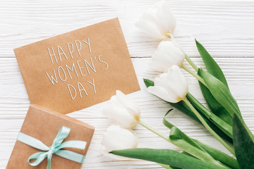 happy womens day text sign on stylish craft present with greeting card and tulips on white wooden rustic background. flat lay with flowers and gift with space for text.  greeting card