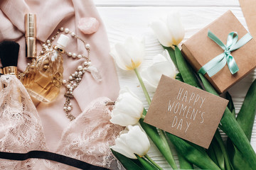 happy womens day text sign on woman lace lingerie jewelry and perfume present on soft fabric and tulips with empty greeting card on white rustic background. flat lay