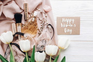 happy womens day text sign on stylish greeting card template with lace lingerie jewelry and perfume on soft fabric and tulips on white rustic background. flat lay